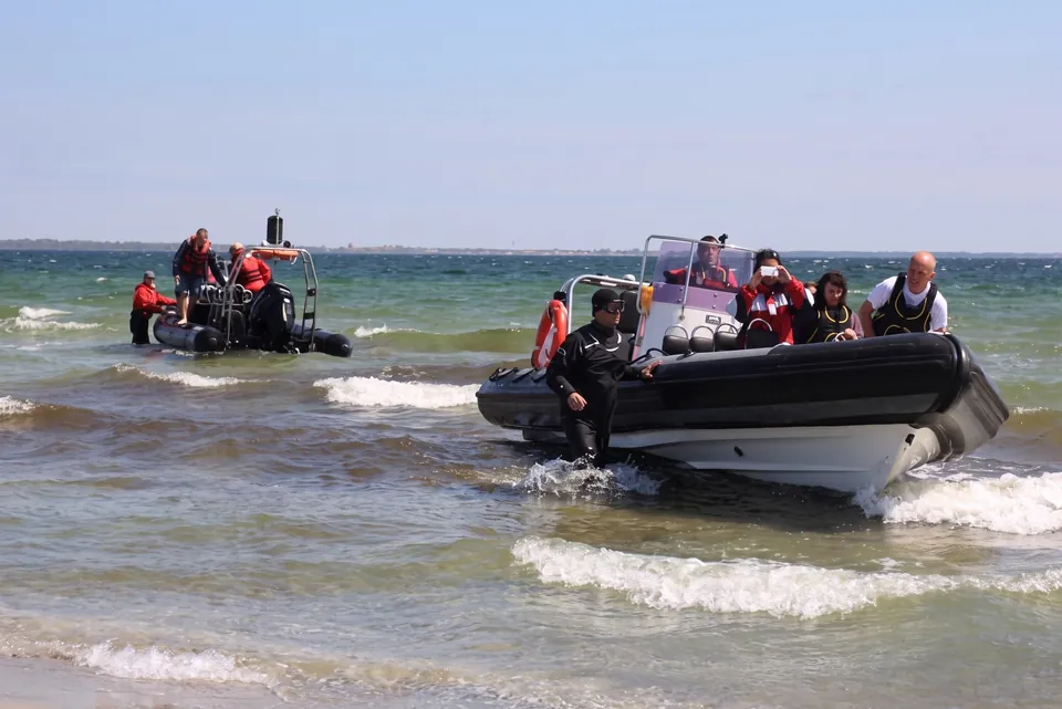 Två ribbåtar med deltagare nära stranden på grunt vatten en solig dag