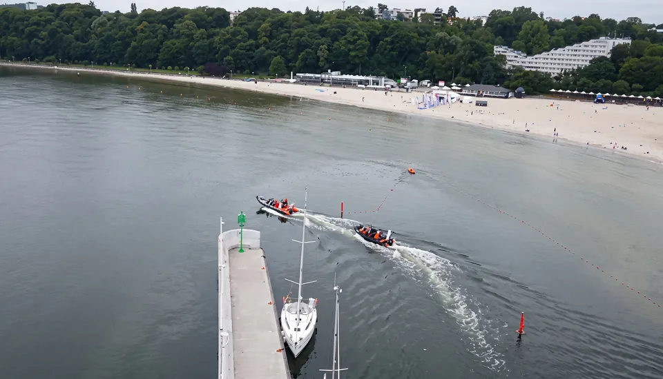 Film still: a RIB boat sailing past a pier and beach, a yacht alongside